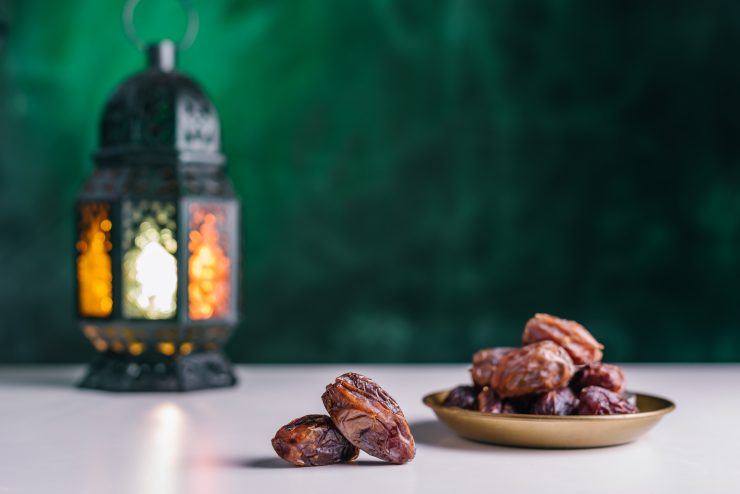 Dates close-up in the foreground. On the distant plan a slightly blurry Burning, lighting, glowing Ramadan Lantern on a white table, textured dark green wall background.