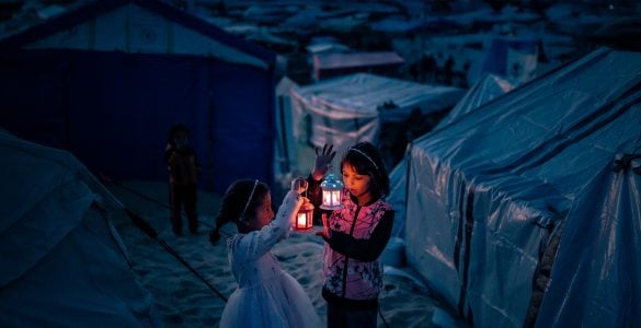 Two young girls in a refugee camp hold glowing lanterns against the backdrop of tents, their faces illuminated with warmth and resilience amidst the darkness. This image reflects the spirit of Gaza in Ramadan—caught between hardship and hope, echoing the soul’s journey between Pharaoh’s oppression and the Divine promise