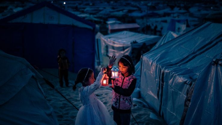 Two young girls in a refugee camp hold glowing lanterns against the backdrop of tents, their faces illuminated with warmth and resilience amidst the darkness. This image reflects the spirit of Gaza in Ramadan—caught between hardship and hope, echoing the soul’s journey between Pharaoh’s oppression and the Divine promise